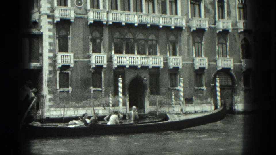 Black-and-white 8mm Venice frame showing passengers in a gondola beside ornate canal-side architecture.