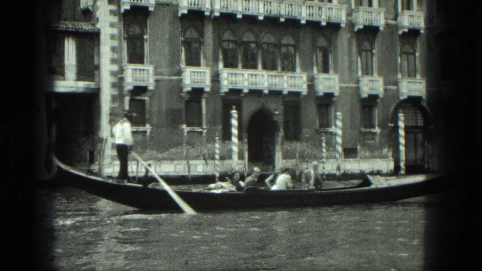 1975 Venice home-movie frame showing a gondolier steering a gondola past ornate canal-side buildings.
