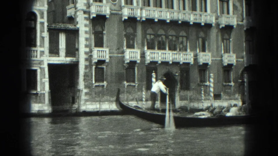 1975 Venice frame with a gondolier using a long oar as the gondola passes arched windows and balconies.