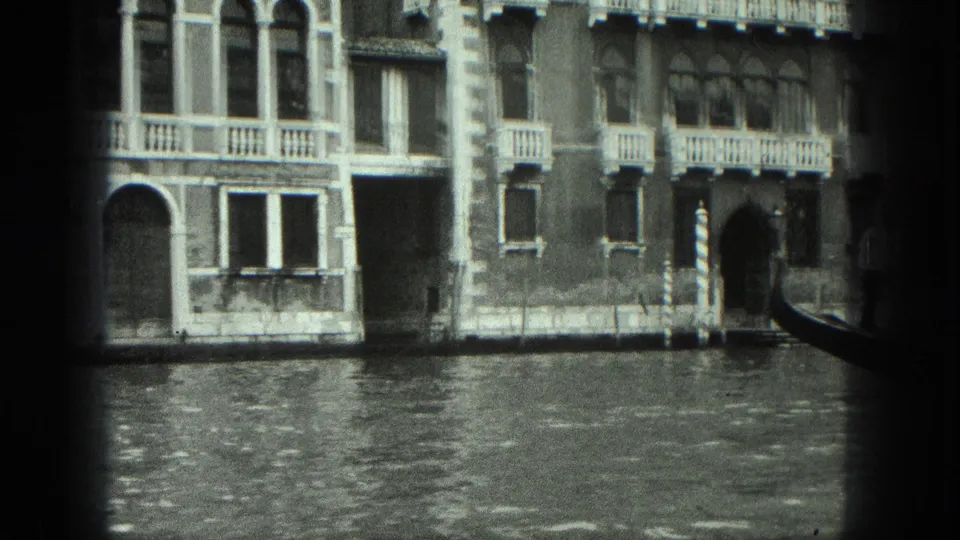 1975 Venice canal view with building windows, balcony rails, water, and part of a gondola at the right edge.