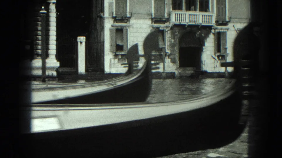 1975 Venice frame showing the curved bow of a gondola crossing in front of canal-side doorways and facades.