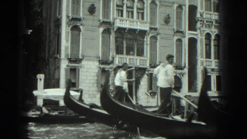 1975 Venice frame with gondoliers, gondola bows, passengers, and multi-story canal-side architecture.