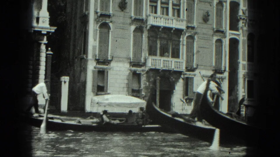 1975 Venice canal view with a gondolier steering past ornate palazzi, balconies, and striped mooring poles.