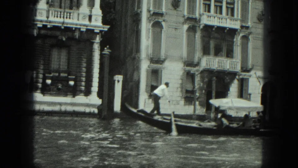 1975 Venice footage shows a gondola carrying a passenger past canal-front buildings and striped mooring posts.