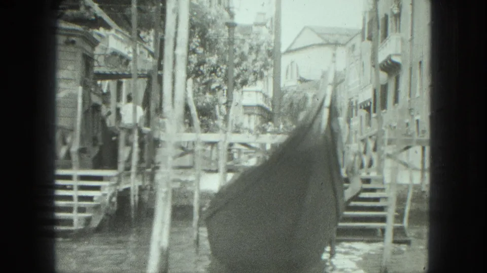1975 Venice canal perspective from near a gondola prow, with buildings, railings, water, and a small bridge-like crossing ahead.