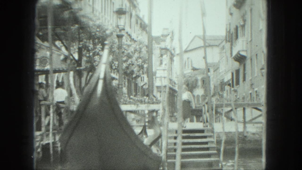 1975 8mm Venice canal view with a gondola bow in the foreground, mooring poles, stone steps, and multi-story buildings along the water.