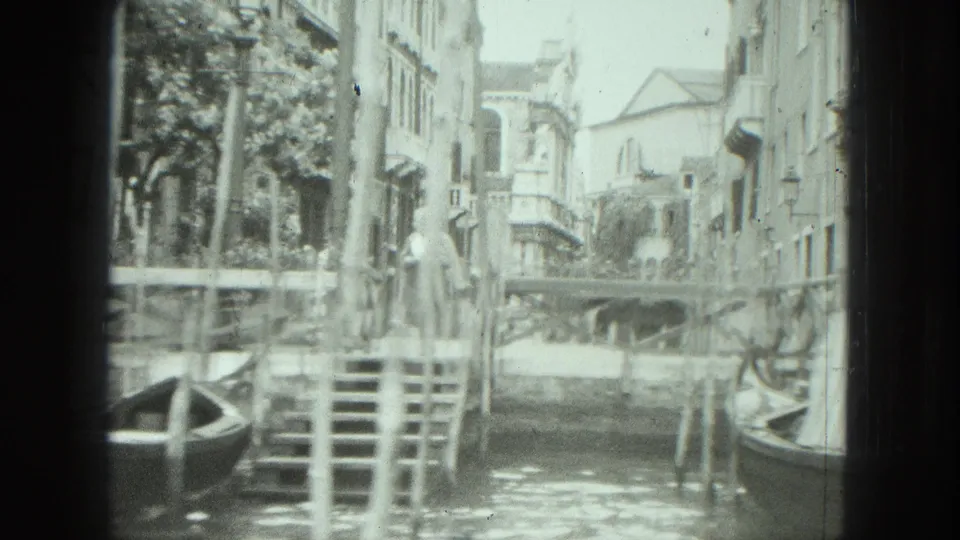 1975 Venice canal scene with a woman standing near stone steps, a gondola at the waterline, mooring poles, and residential facades behind her.