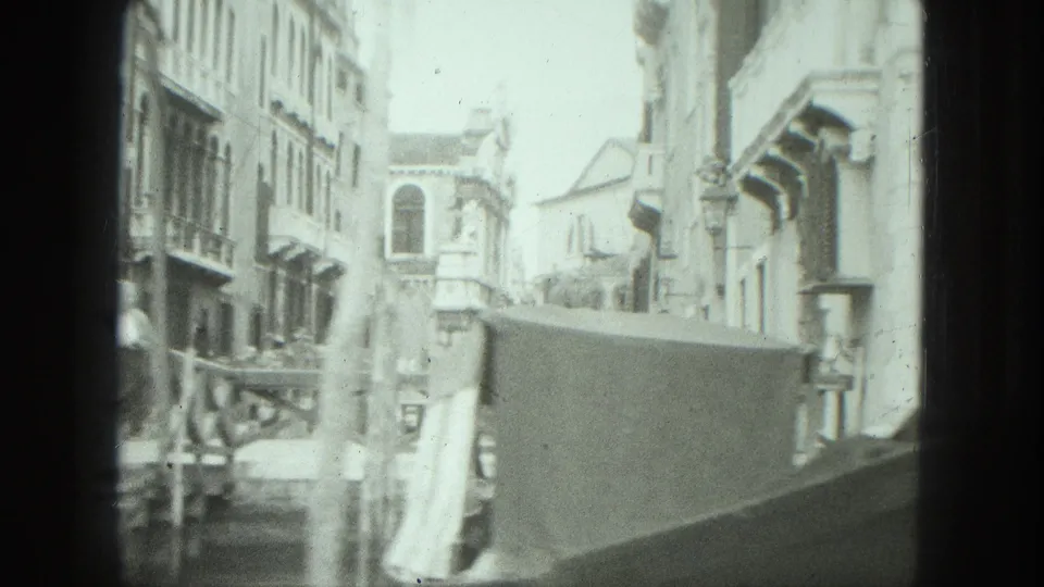 Black-and-white 8mm view along a Venice canal, showing a gondola edge, stone walls, windows, balconies, and pale building facades.