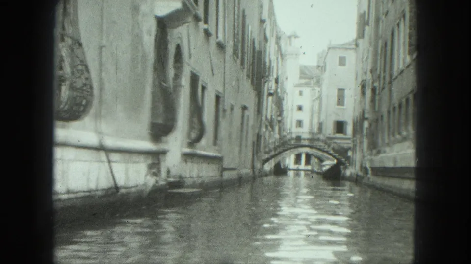 1975 Venice canal view looking toward a small arched bridge, with tall waterside buildings, windows, and rippled water in black-and-white 8mm footage.