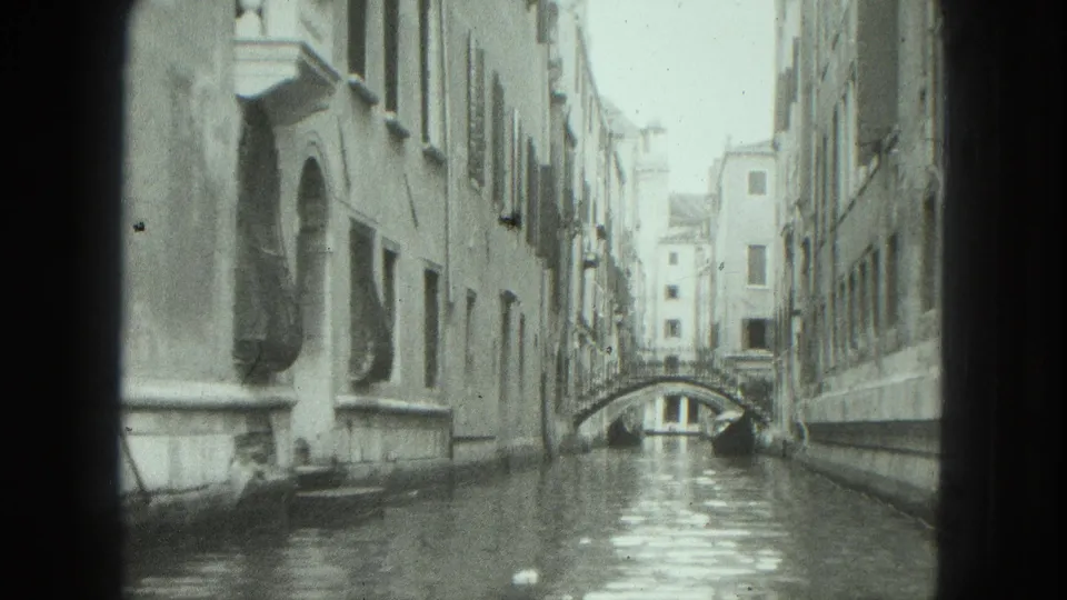 1975 8mm view of a narrow Venice canal lined with weathered buildings and crossed by a small arched bridge.