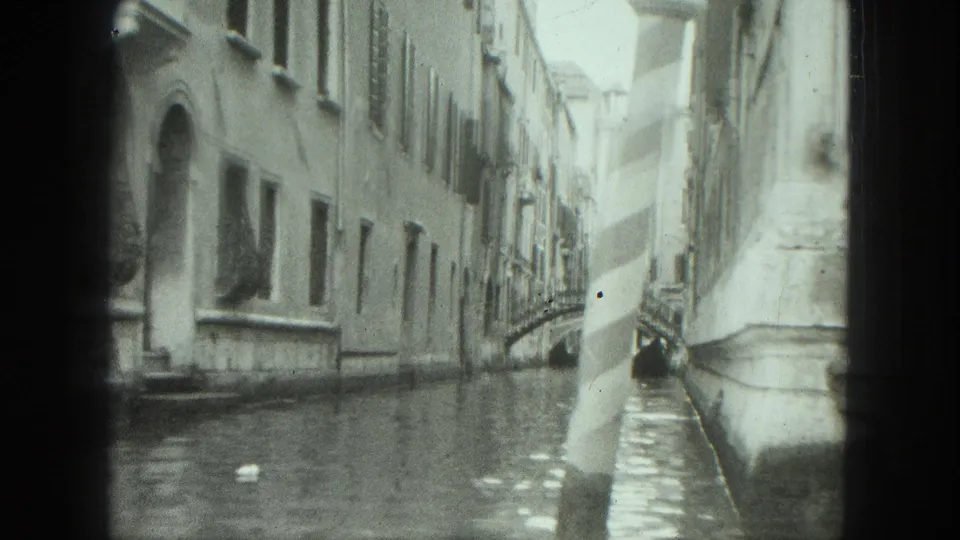 Venice canal scene on 8mm film, showing pale building facades, calm water, and an arched bridge in the distance.