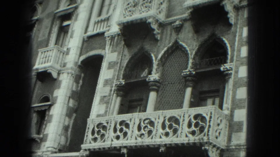 8mm close-up of a Venice building facade with ornate balcony stonework, pointed arches, and textured masonry.