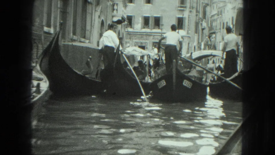 1975 Venice canal view with gondoliers standing on gondolas, passengers nearby, and historic buildings lining the waterway.