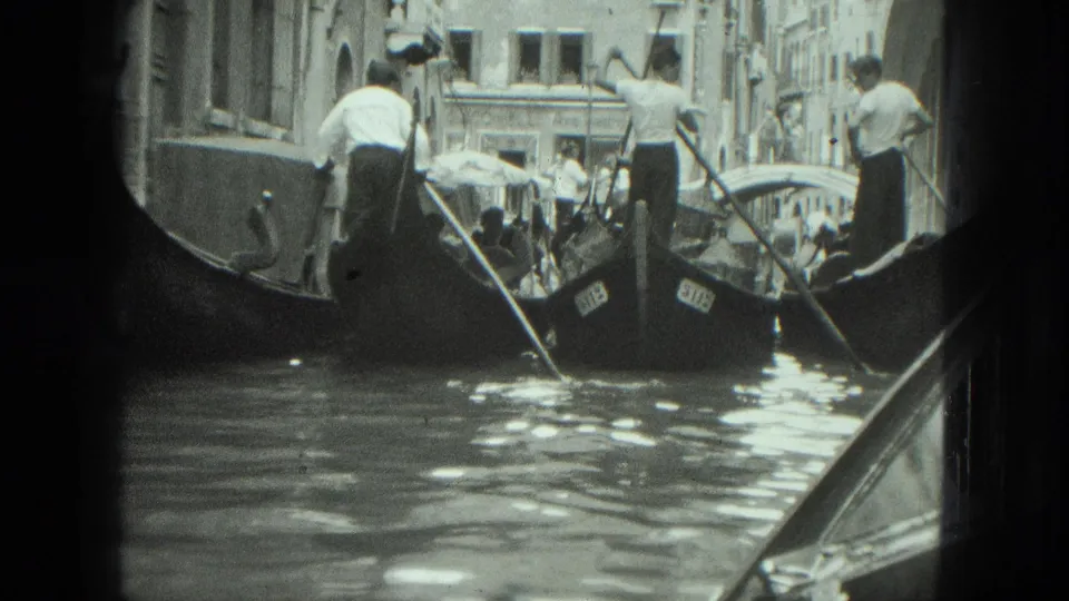 1975 Venice footage frame showing multiple gondolas, standing gondoliers, passengers under a canopy, and a small bridge ahead.