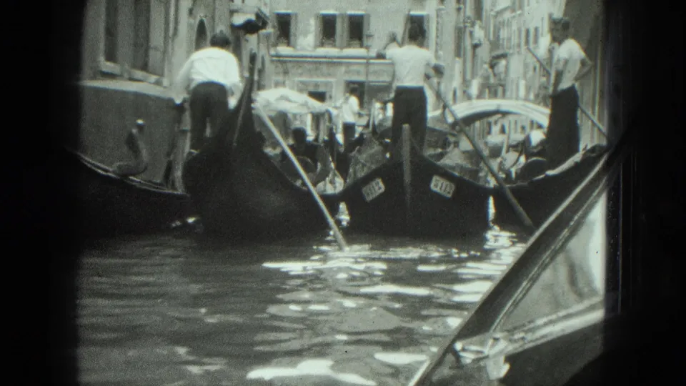 1975 Venice canal frame from a boat-level perspective, showing gondolas, gondoliers, passengers, rippled water, and narrow buildings.