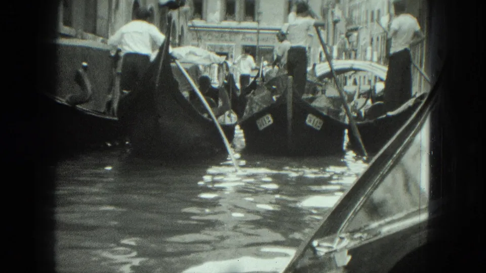 1975 Venice canal scene with gondoliers standing at the sterns of tour boats as passengers ride beneath canvas covers.