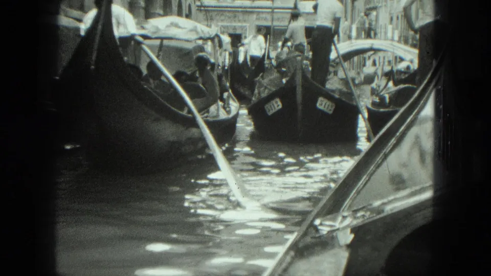 1975 home-movie view of gondoliers and passengers in Venice canal traffic, with water reflections and architecture behind them.