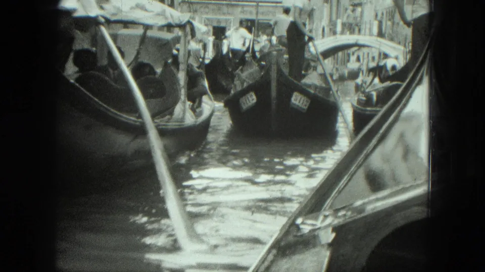 Venice canal traffic in 1975 with gondoliers standing over seated passengers as another gondola edge fills the foreground.