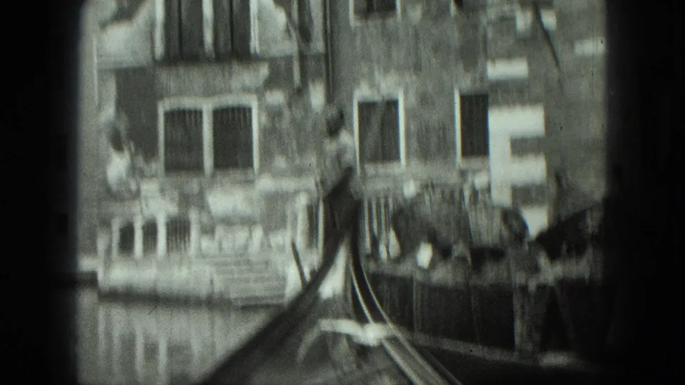Soft black-and-white 8mm frame from Venice showing a gondola bow, canal water, weathered facade, windows, and balcony details.