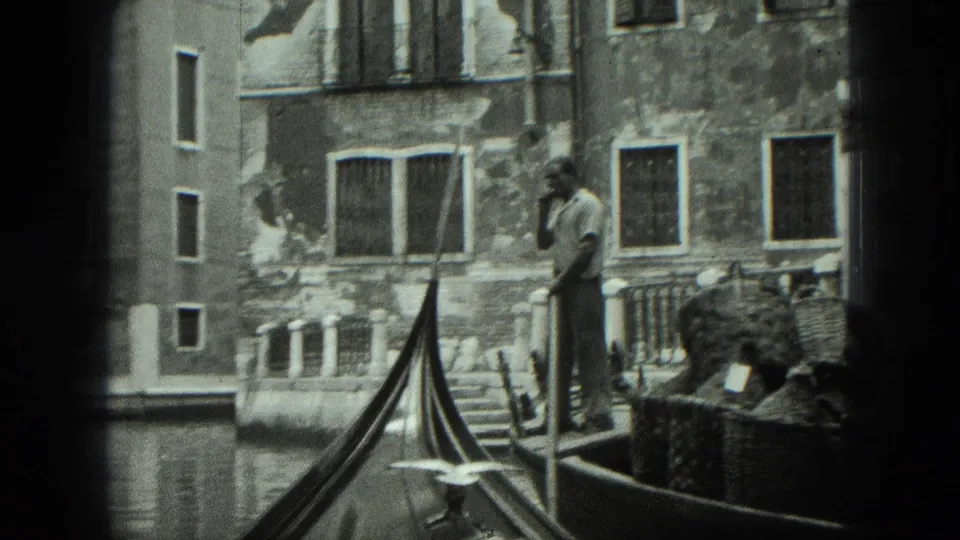 1975 Venice home-movie frame with a standing gondolier, long oar, gondola bow, canal water, old facades, and window grilles.