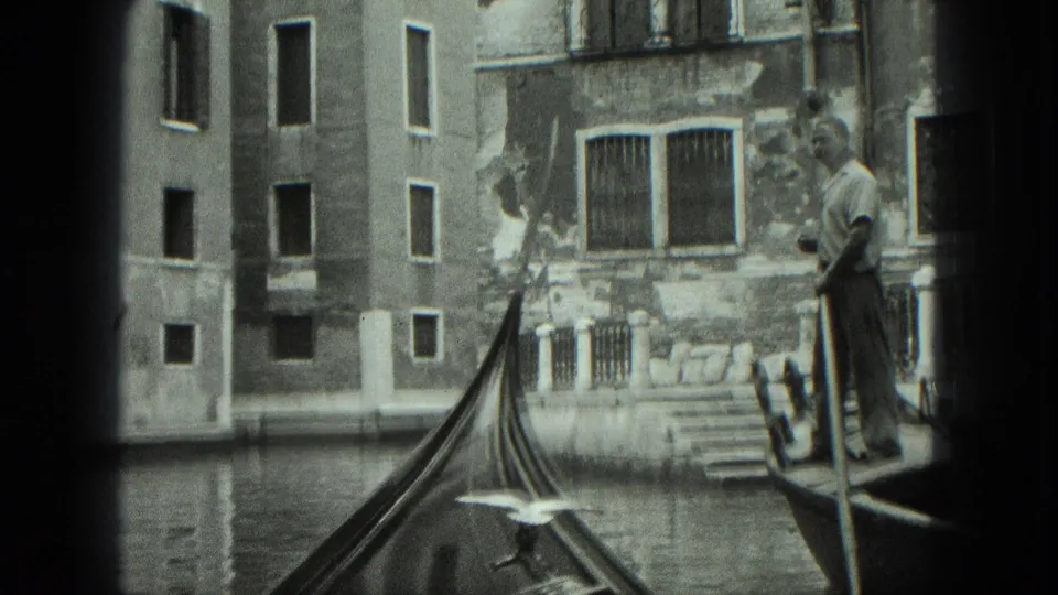 Venice 1975 black-and-white 8mm frame showing a gondolier, pointed gondola bow, calm canal water, and weathered residential facades.