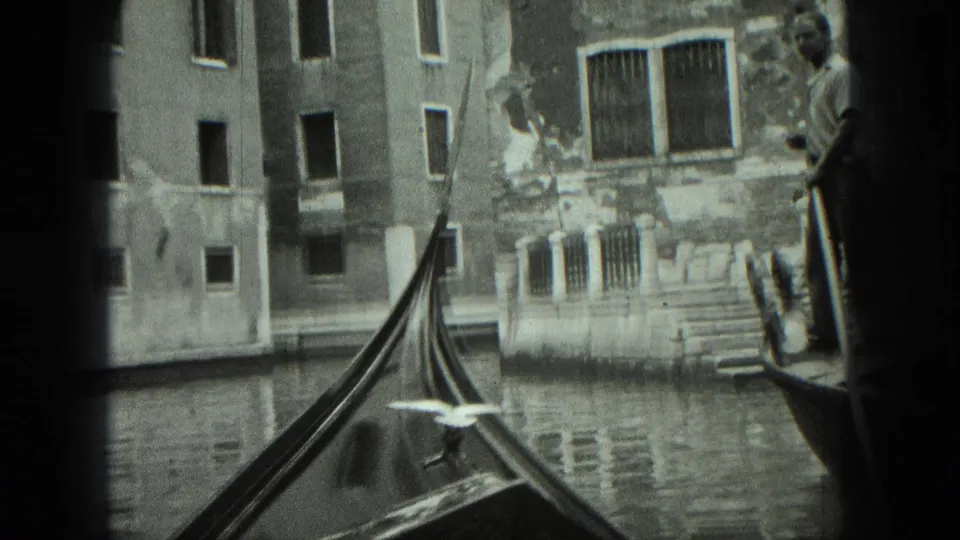 1975 8mm Venice gondola view with the boat prow, a gondolier, canal water, and weathered buildings along the waterway.
