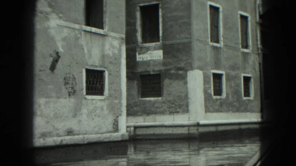 1975 8mm Venice canal view showing a weathered stucco corner building, barred windows, and calm canal water.