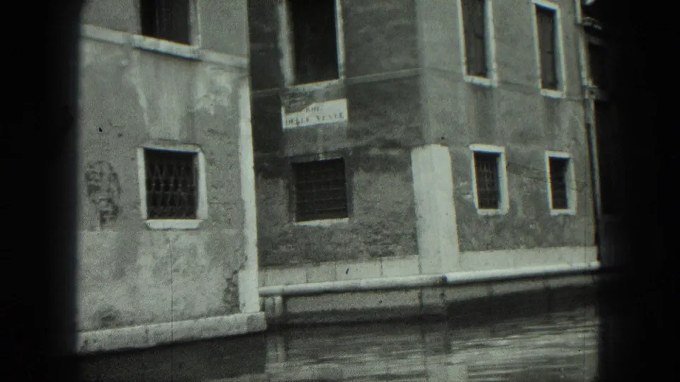 1975 8mm Venice canal shot with a stucco wall, barred lower windows, an open upper window, and waterline stonework.