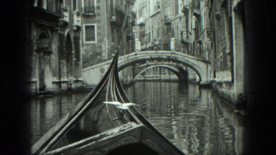 1975 8mm Venice gondola perspective looking toward a stone arched bridge between tall canal-side buildings.