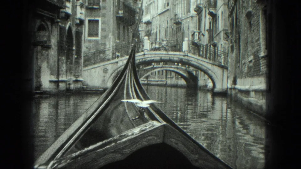 1975 Venice 8mm view from a gondola, showing the prow, canal water, old buildings, and an arched stone bridge ahead.