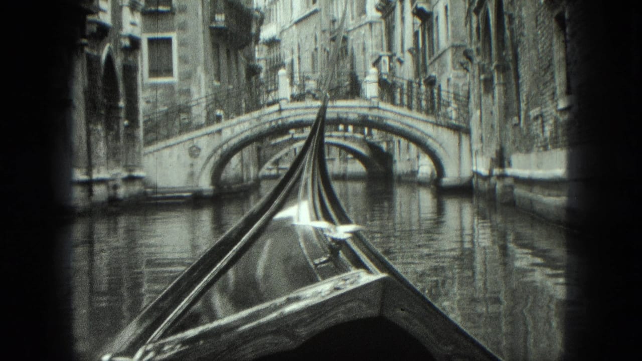 Gondola bow centered in a canal with a stone bridge and buildings ahead.
