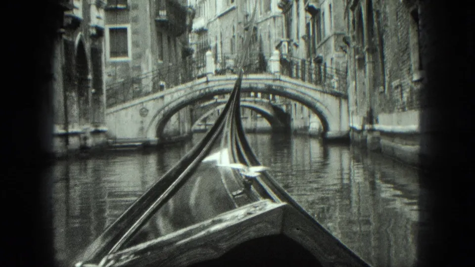 Black-and-white 8mm Venice canal view from a gondola, with the bow aligned toward a small arched bridge and surrounding buildings.