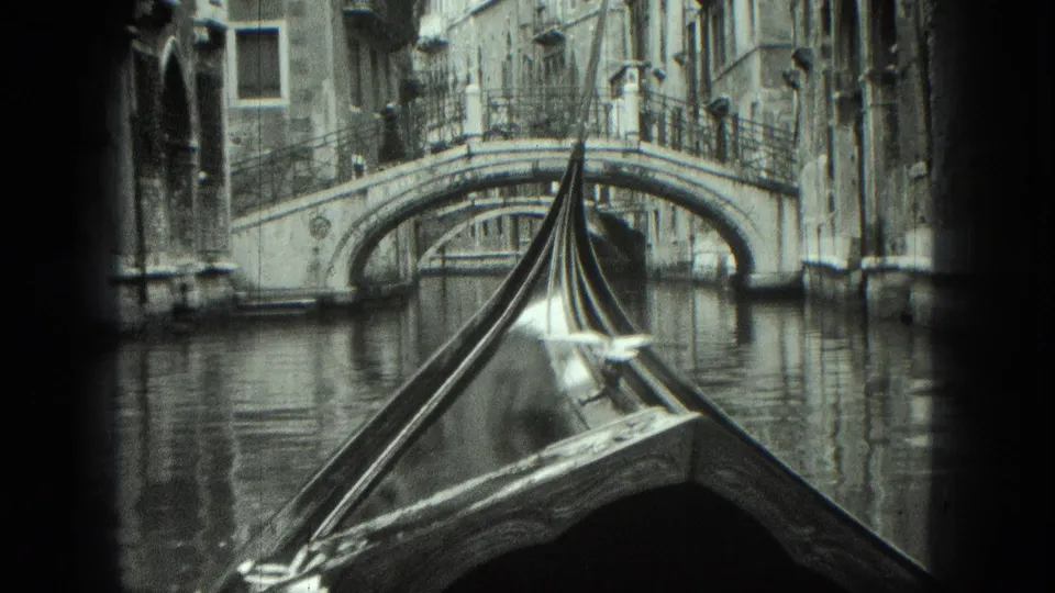 1975 Venice home-movie frame showing a gondola prow close to a stone bridge, with still canal water and old building fronts around it.