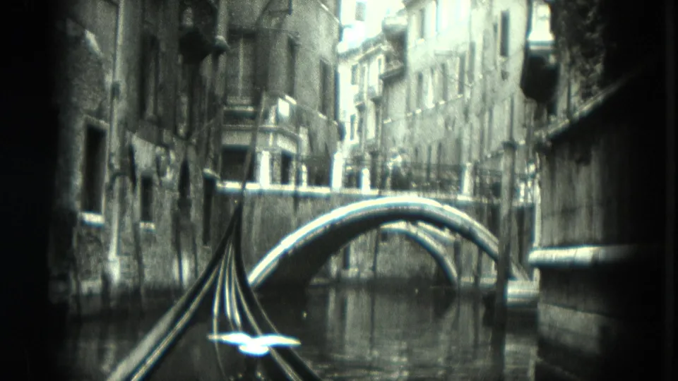 1975 8mm Venice canal view from a gondola, with stone buildings, an arched bridge ahead, reflective water, and gondola fittings in front.