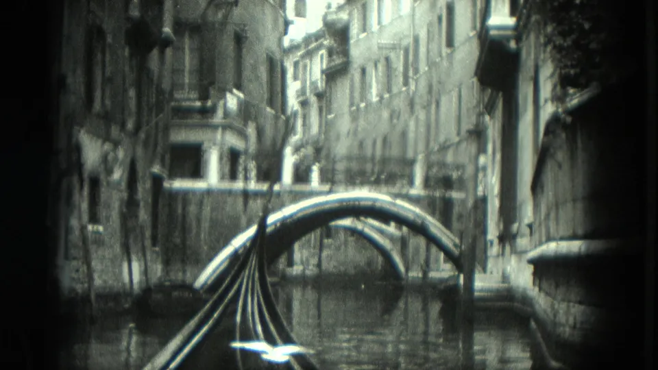 Black-and-white 8mm Venice gondola view showing a narrow canal, an arched bridge, aged building facades, and rippling reflections.