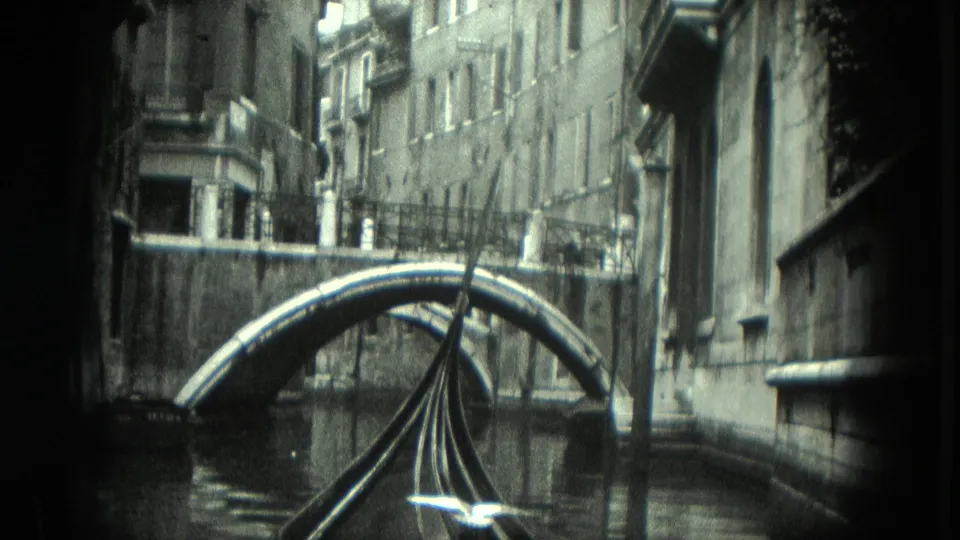 The gondola viewpoint nears a curved bridge in Venice, with weathered building facades and canal reflections around it.