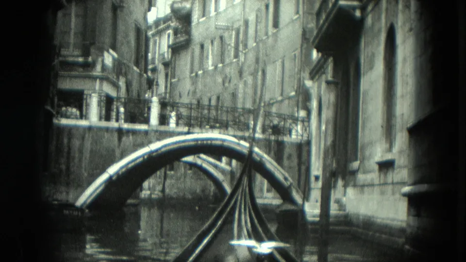 A close Venice canal frame shows the gondola prow aligned with the bridge, flanked by masonry walls and windows.