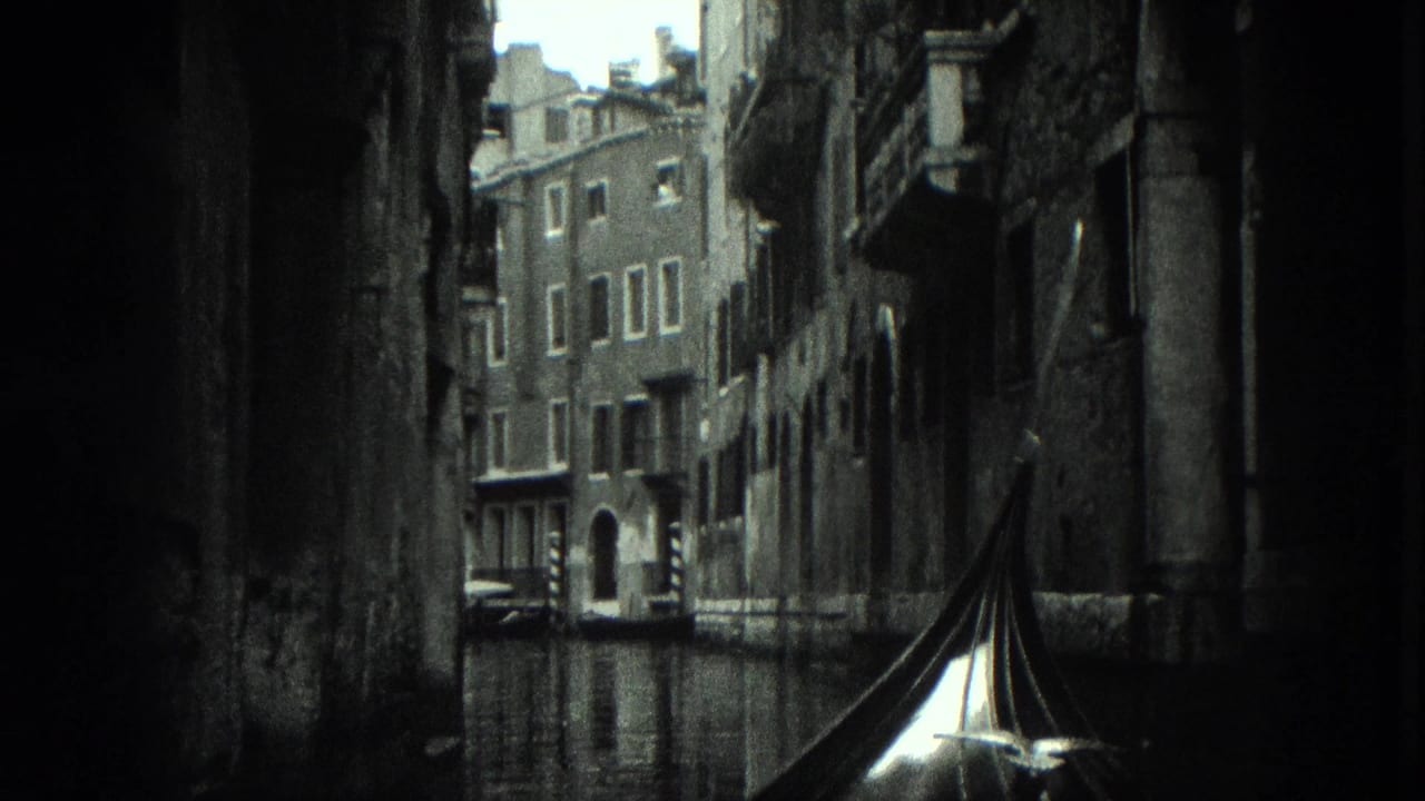 Black-and-white 1975 8mm view from a gondola traveling through a narrow Venice canal lined with weathered masonry buildings.