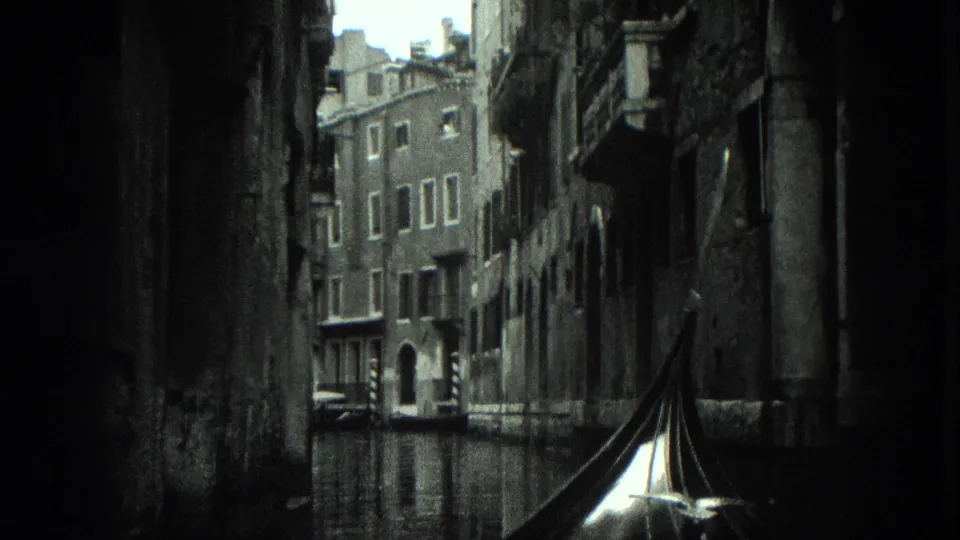 Black-and-white 1975 8mm view from a gondola traveling through a narrow Venice canal lined with weathered masonry buildings.