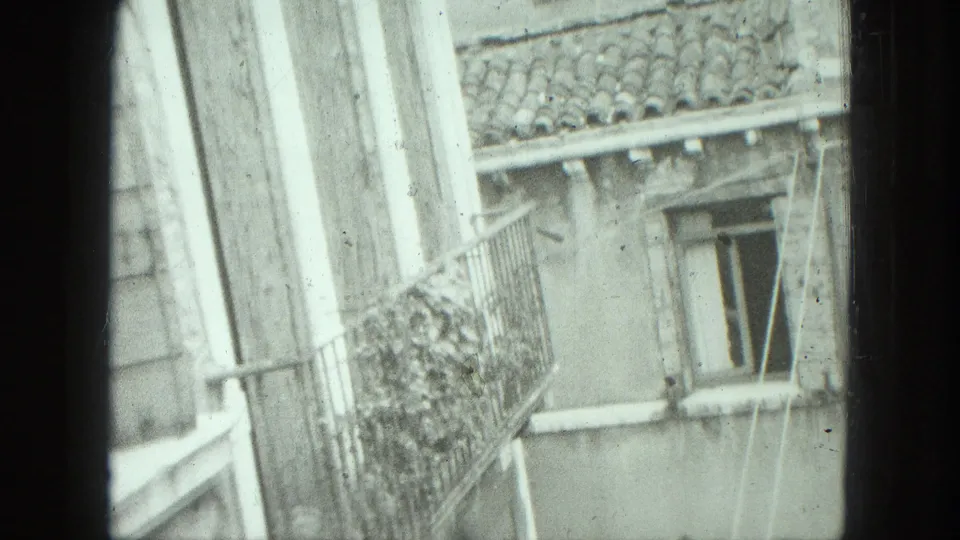 1975 Venice home-movie frame with an angled facade view, balcony plants, roof tiles, and a small window near the edge of frame.