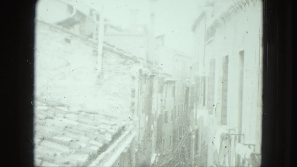 1975 Venice frame looking across roof tiles toward a tall facade with shuttered openings and a small balcony rail.