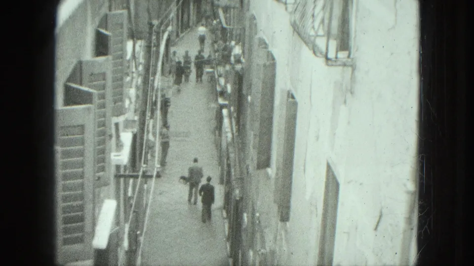 Elevated 1975 Venice home-movie frame of pedestrians walking through a tight street bordered by pale walls and shuttered windows.