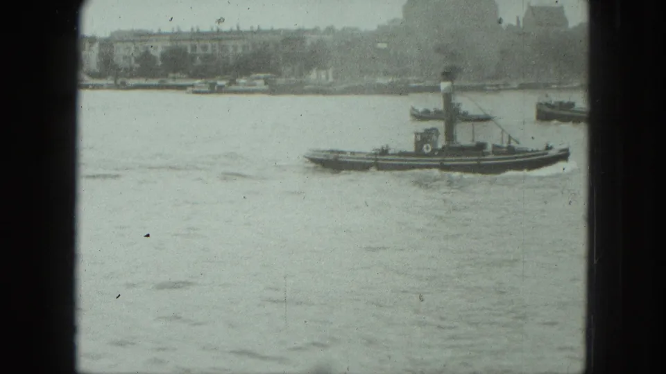 1975 Venice harbor frame showing a small tugboat crossing calm water near distant waterfront buildings.