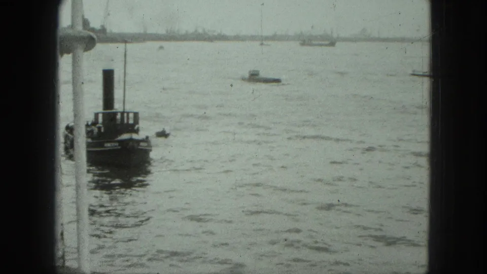 A 1975 Venice home-movie frame looks past a white window edge toward a dark harbor boat and several small distant vessels.