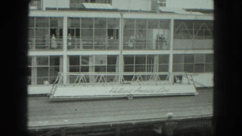 Venice port scene showing a glass-fronted terminal and a gangway marked Holland America Line, with people visible inside.