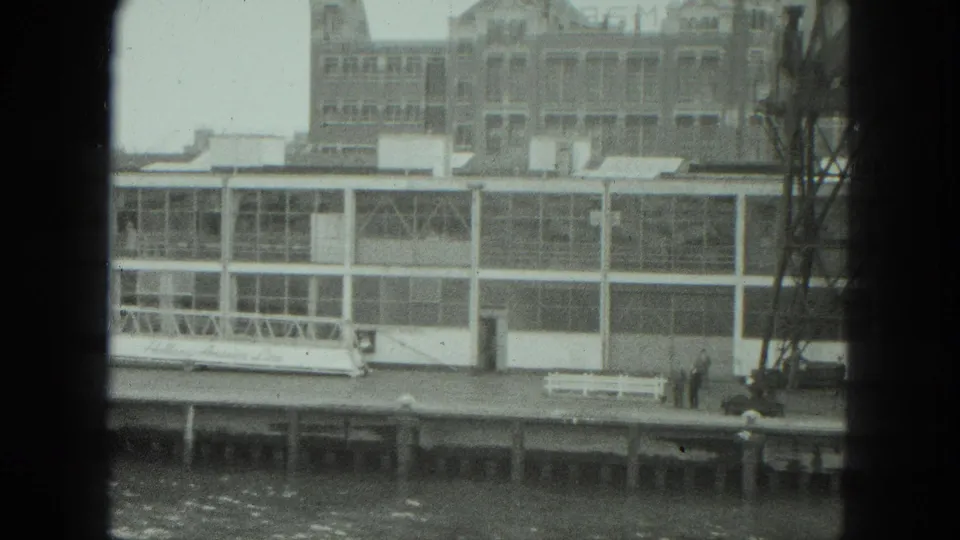 1975 Venice waterfront frame showing a white dock structure, bench, distant people, and a metal tower at right.