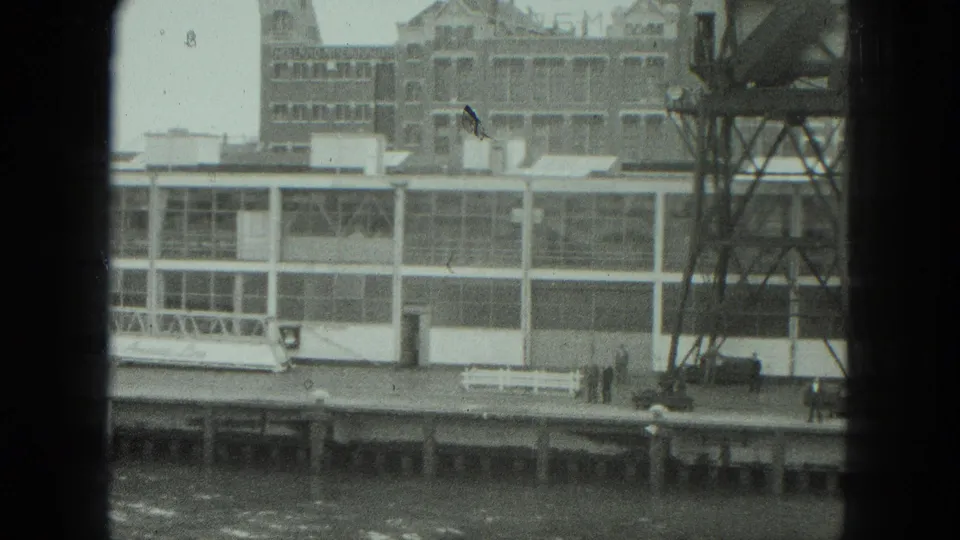 1975 Venice dockside view with a metal tower, white bench, distant people, and glass-walled waterfront structure.