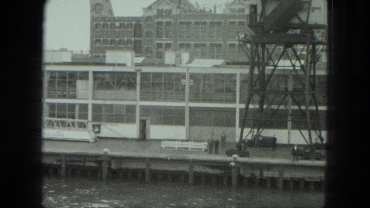 1975 8mm view of a Venice industrial riverbank with a long factory facade, metal tower, pier, bench, and small distant figures.