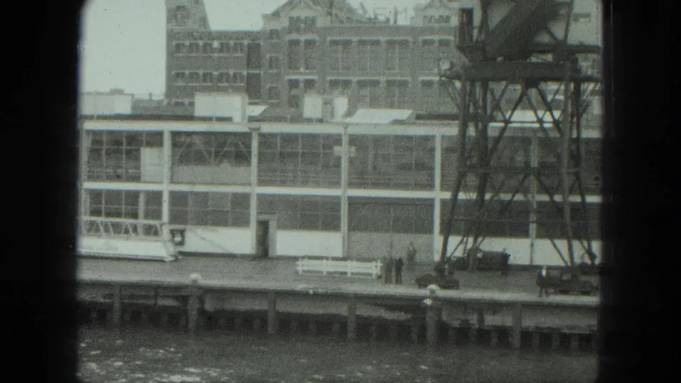 1975 8mm view of a Venice industrial riverbank with a long factory facade, metal tower, pier, bench, and small distant figures.
