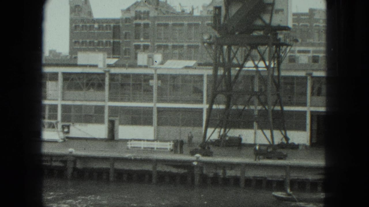 Factory building and metal tower beside a dock, with small figures near the river.